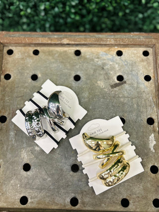 Two sets of metallic sculpted hair tie cuffs on white packaging cards displayed on perforated metal surface.