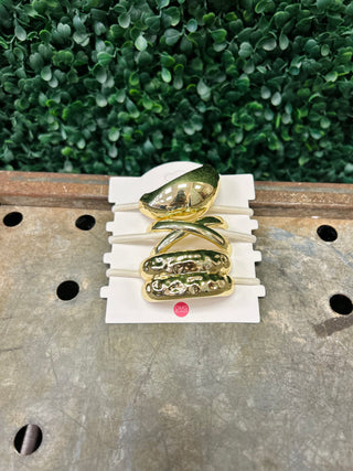 Set of gold-toned sculpted metal hair tie cuffs displayed on white card against wooden surface.