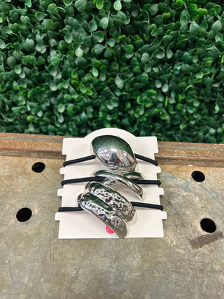 Three silver metal hair ties with sculptural designs on elastic bands displayed on white card against wooden surface.