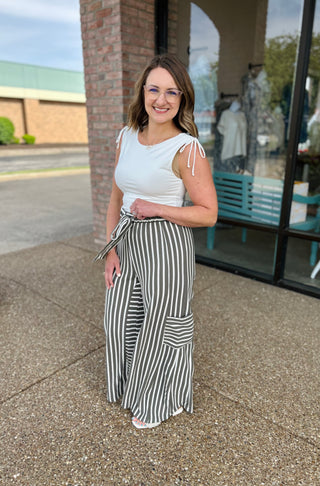 Woman wearing white tie-shoulder top with striped wide-leg pants, standing outdoors near brick storefront.