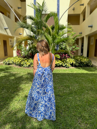 Woman in blue floral backless maxi dress standing on resort lawn with tropical plants and buildings.