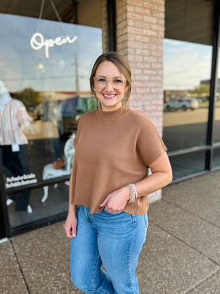 Woman wearing a ribbed mock neck short sleeve sweater top, paired with jeans, outside a boutique.