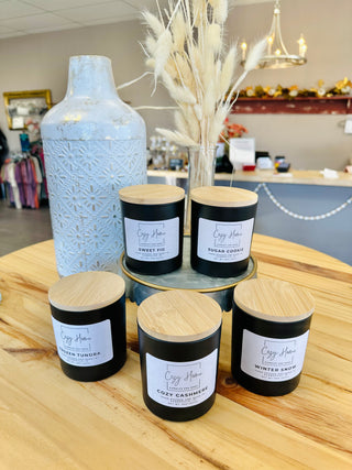 Collection of black soy candles with wooden lids and white labels displayed on wooden table with decorative vase.