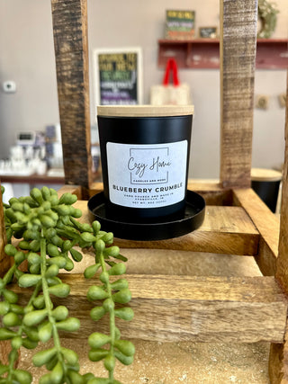 Black soy candle with wooden wick and lid displayed on rustic wooden shelf with succulent plant.