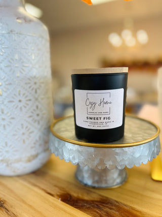Black soy candle with wooden lid displayed on decorative glass cake stand with white vase in background.