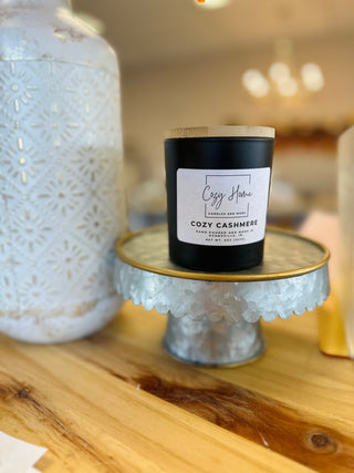 Black soy candle with wooden lid displayed on decorative glass cake stand with white ceramic vase nearby.