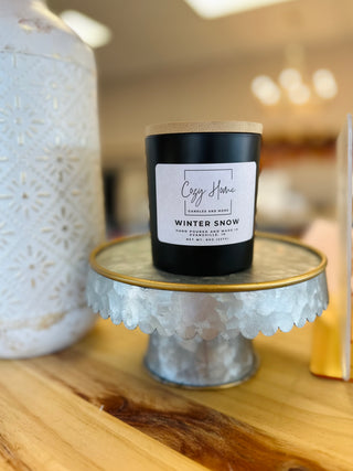 Black soy candle with wooden wick displayed on decorative glass cake stand in cozy home setting.