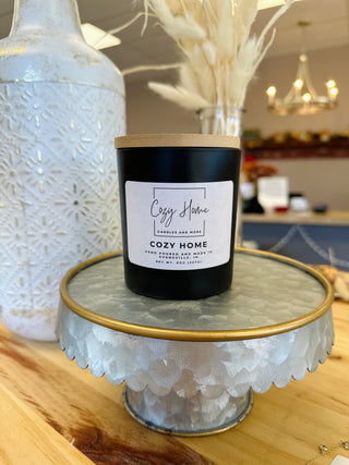 Black soy candle with wooden wick displayed on decorative glass cake stand in cozy home setting.