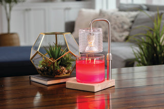 Silver candle warmer lamp melting red wax in glass jar on wooden table with decorative plants