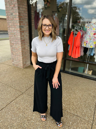 Woman wearing silver ribbed short-sleeve bodysuit with black wide-leg pants, standing outside storefront.