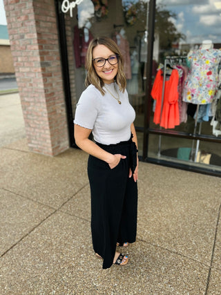 Woman wearing silver ribbed short-sleeve bodysuit with black pants standing outside clothing boutique storefront.