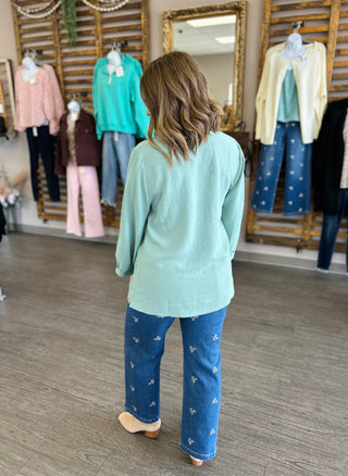 Woman wearing mint green long-sleeve top and embroidered jeans in clothing boutique setting.