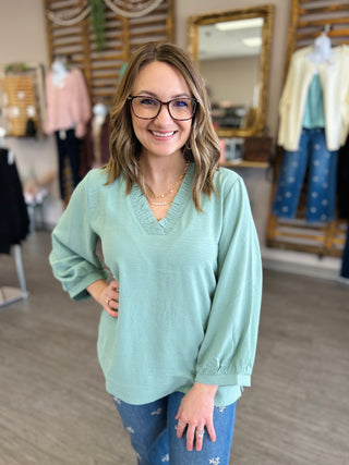 Woman wearing mint green v-neck sweater with layered gold necklaces in clothing boutique setting