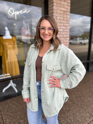 Woman smiling outside boutique wearing an open ribbed knit shacket over brown top and jeans.