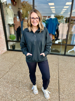 Woman wearing gray quarter zip pullover with black pants and sneakers, standing outside retail storefront.