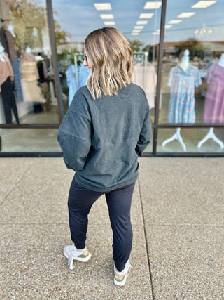 Woman wearing gray quarter zip pullover and dark pants, standing outside clothing store window display.