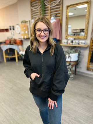 Woman wearing dark zip-up hoodie with textured fabric and kangaroo pocket, standing in retail store.