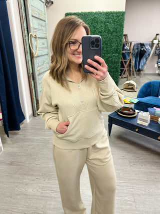 Woman wearing beige ribbed knit matching set with half-zip top and pants in retail store mirror selfie.