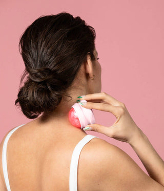 Woman using heart-shaped massage tool on neck and shoulder area against pink background.