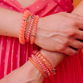 Two hands wearing coral and pink spiral hair coils as bracelets against vibrant pink clothing.