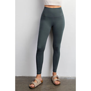 Woman wearing high-waisted athletic leggings with white crop top and neutral sandals against gray wall.