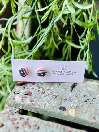 Star-spangled stud earrings on Harper Klay Co. card displayed on weathered wood with green foliage background.