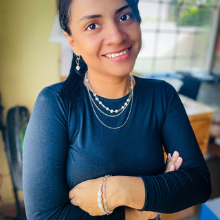 Woman wearing silver layered necklaces and pearl bracelet, smiling in casual navy top indoors.