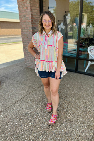 Woman wearing colorful striped sleeveless top with tiered design and pink trim detailing, styled with denim shorts.