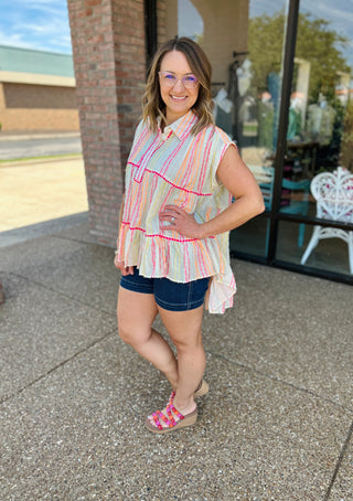 Woman wearing colorful striped sleeveless top with pink trim detail and denim shorts, standing outdoors.