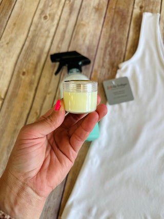 Hand holding small jar of yellow stain remover with spray bottle and white cloth on wooden background.