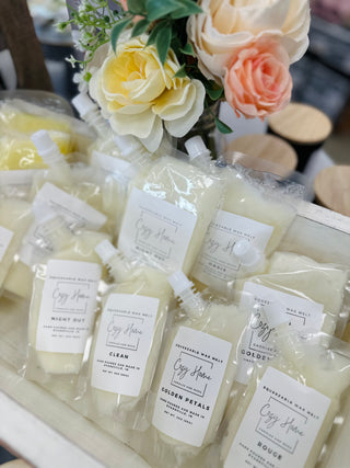Squeezable wax melts in clear packages with white labels displayed alongside decorative flowers and candles.