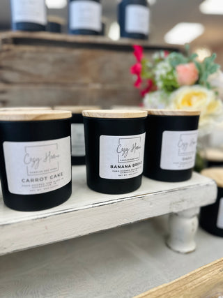 Three black soy candles with wooden lids and white labels displayed on wooden shelf in kitchen setting.
