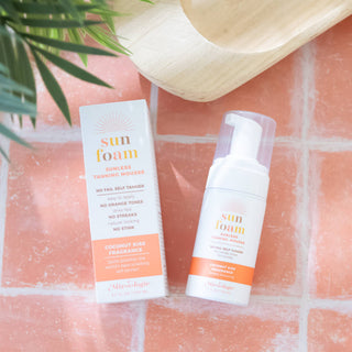 SunFoam sunless tanning mousse bottle and box displayed on coral tile surface with palm leaf and woven basket.