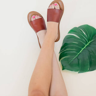 Woman's legs wearing brown leather slide sandals with pink pedicure next to tropical leaf