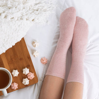 Person wearing soft pink spa socks relaxing in bed with coffee and meringue treats on wooden tray.