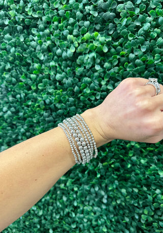 Woman wearing multiple sparkling beaded stretch bracelets and ring against green foliage background.
