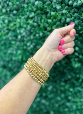 Hand wearing multiple gold beaded stretch bracelets with bright pink nails against green foliage background.