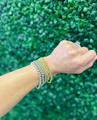 Hand wearing multiple gold and silver beaded stretch bracelets with matching ring against green foliage background.