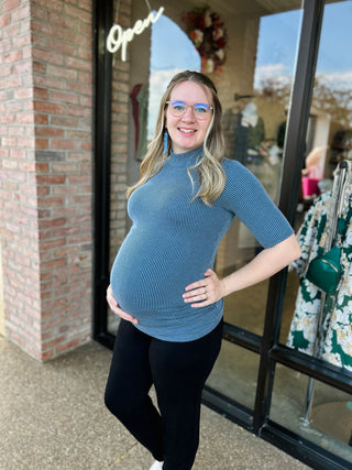 Pregnant woman wearing blue ribbed mock neck maternity top, standing outside storefront with hand on hip.