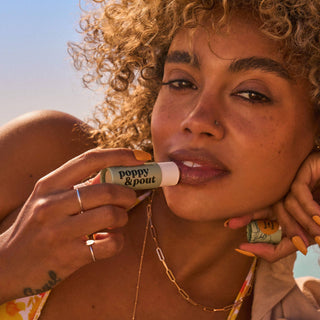 Woman applying Poppy & Pout Sweet Mint Lip Balm in golden sunlight with curly hair and jewelry.