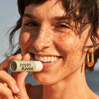 Smiling woman with freckles holding Poppy & Pout Sweet Mint Lip Balm tube near her face in sunlight.