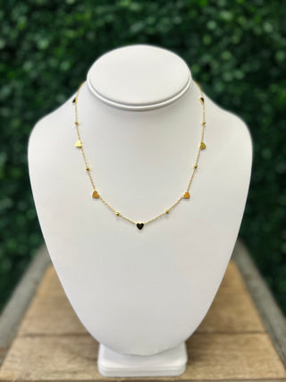 Gold heart charm necklace displayed on white jewelry bust stand with green foliage background