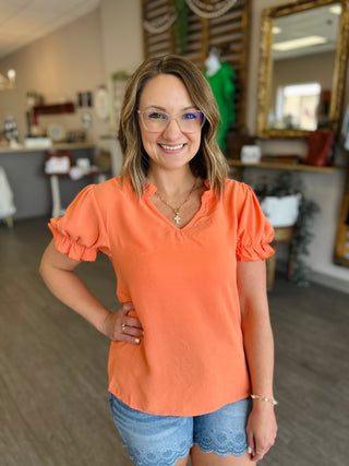 Woman wearing orange ruffled sleeve v-neck top with denim shorts, standing in bright interior space.