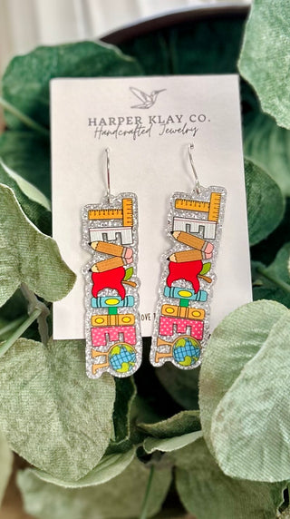 Colorful school-themed glitter earrings with stacked educational supplies design on product card with green leaves.