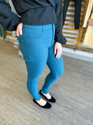 Woman wearing teal stretch skinny pants with dark jacket and black flats on wooden floor.