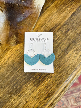 Handcrafted clay hoop earrings with textured teal pendants displayed on branded card against wooden surface.