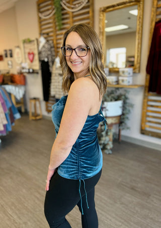 Woman wearing teal velvet tank top in boutique store, smiling over shoulder at camera.
