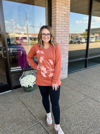Woman wearing terracotta hoodie with floral design standing outside brick storefront, smiling at camera.
