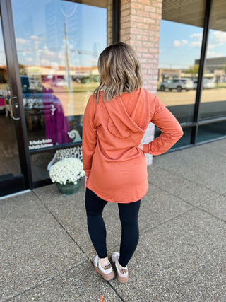 Woman wearing Rae Mode Terracotta Sunset Hoodie with leggings and sneakers, standing outside storefront