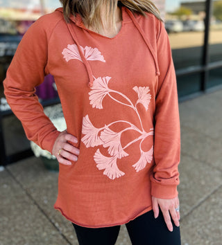 Woman wearing coral terracotta hoodie with pink floral design, standing outdoors with hand on hip.